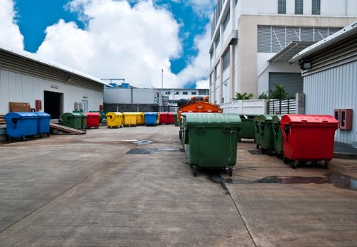 The image shows a paved outdoor area between two industrial or commercial buildings, used for rubbish disposal. In the foreground, multiple large wheelie bins are arranged, with some in green, red, and yellow colours, each featuring a textured plastic surface and metal lids. A few bins are slightly open or tilted, indicating recent use. In the background, a row of additional bins in similar colours extends towards the buildings, which have metal siding and visible ventilation systems. The sky overhead is partly cloudy with patches of blue, suggesting daytime in a typical urban setting. This scene exemplifies the type of rubbish disposal area managed by a local waste removal service such as Office Clearance Greenwich, serving areas including Greenwich and the surrounding postcode districts. The overall environment is neat and organized, with the bins positioned properly on the concrete surface, ready for collection or disposal. The setting highlights typical commercial waste management practices in a city-centre environment, relevant to the services offered by local rubbish removal providers.