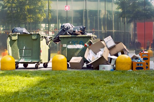 Two green wheelie bins filled with rubbish are positioned on a paved area in front of a reflective glass building, with black garbage bags placed on top of the bins. Several cardboard boxes and other waste items are stacked around the bins, some leaning against them. Bright yellow concrete bollards are situated along the edge of a grassy area in the foreground. The background features the glass facade of a modern office building, reflecting trees and a red object, possibly an emergency or informational sign, on the street side. This scene depicts waste awaiting collection or removal, suitable for a professional rubbish removal service such as Office Clearance Greenwich serving the Greenwich area.