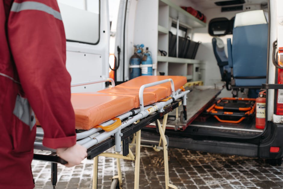 A person in a red uniform is loading an orange cushioned stretcher with metal handles into an ambulance vehicle. The ambulance interior features white walls and shelves, with equipment and supplies vi