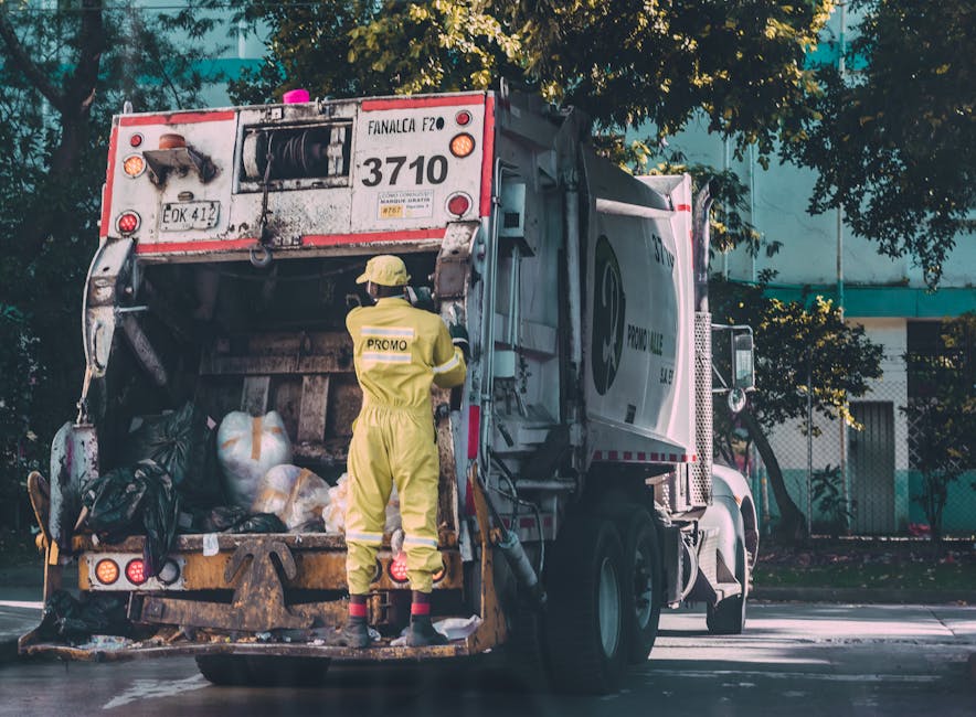 A waste collection vehicle parked on a street during daytime with its rear compartment open, revealing black and white rubbish bags, some tied with orange and white string, and miscellaneous waste mat